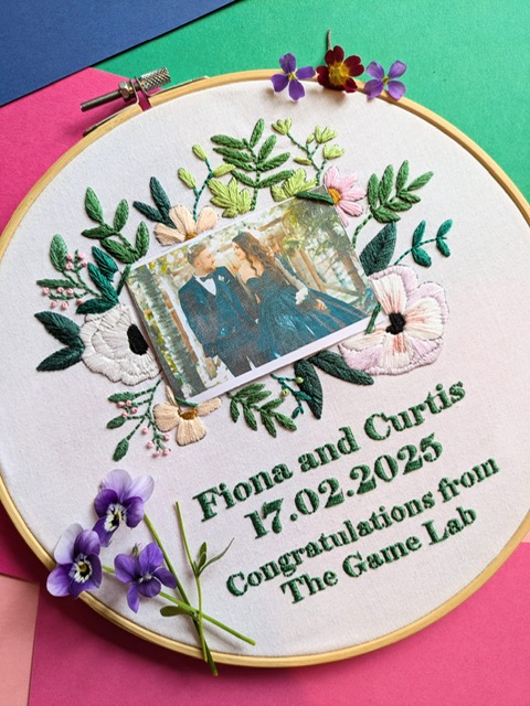 Hand-embroidered hoop saying congratulations from the Game Lab with flowers framing a couple. 
